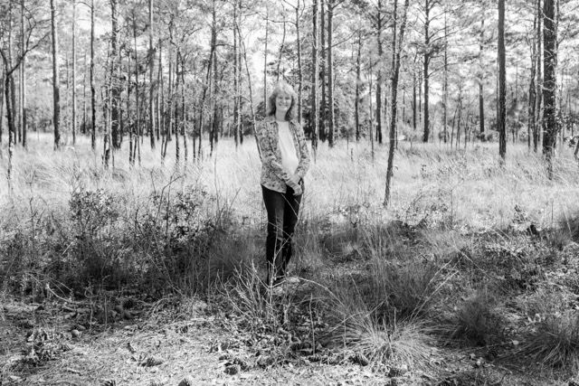 Black and white photo of a woman standing in a pine forest with tall grass.