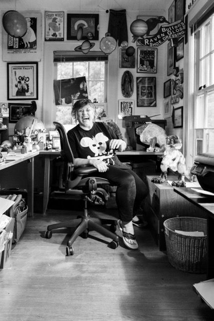 Woman laughing in cluttered art studio office chair, surrounded by posters and collectibles, wearing Mickey Mouse sweater.