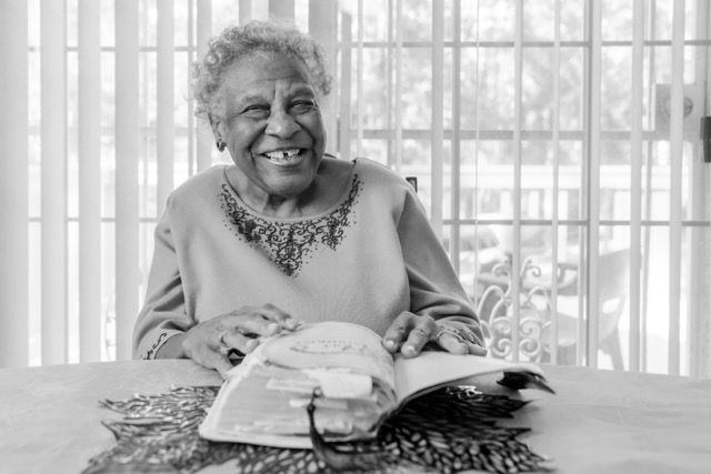 Elderly woman smiling warmly while reading a book at a table near bright window.