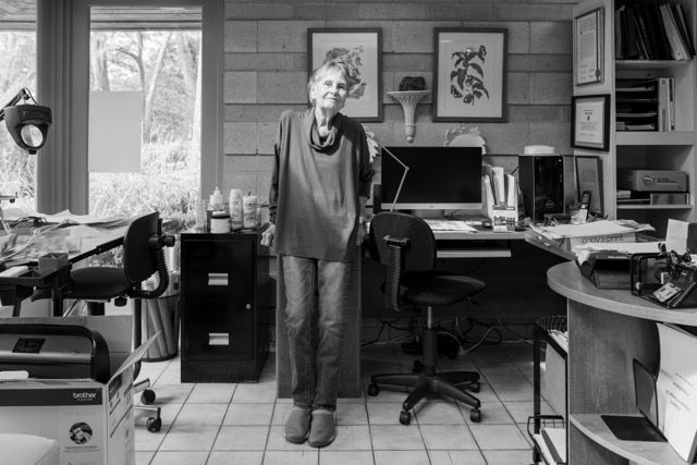 Woman standing in a studio workspace with desk and artwork in black and white.