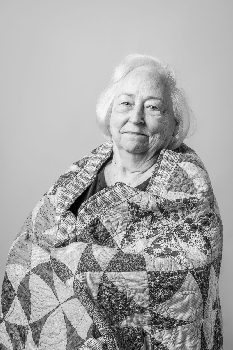 Black and white portrait of a smiling elderly woman with white hair wrapped in a patchwork quilt.