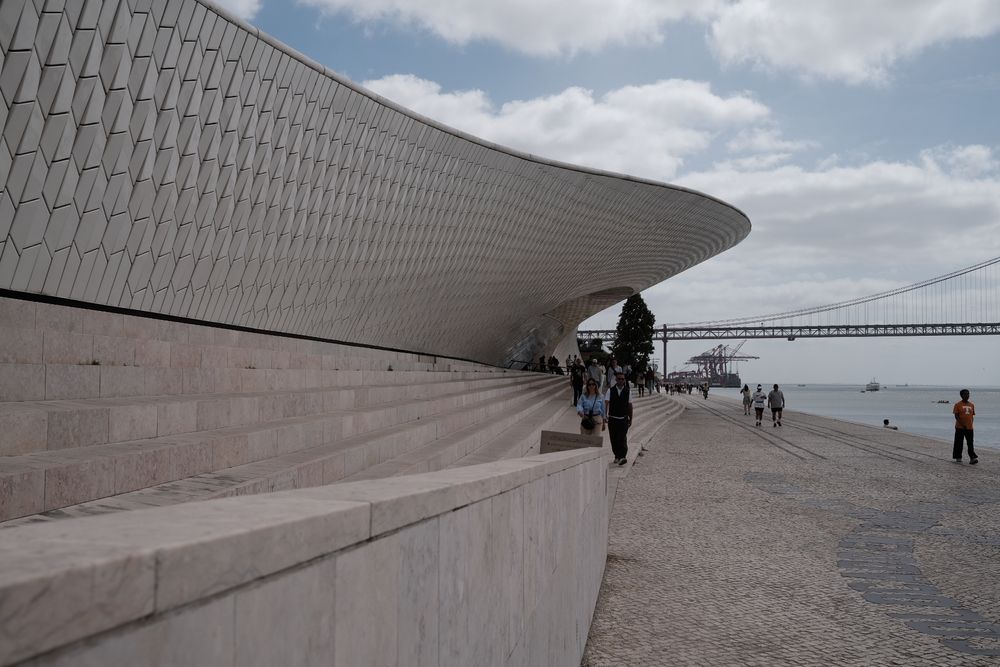 Modern MAAT museum facade in Lisbon with wooden promenade, visitors, and Ponte 25 de Abril bridge over Tagus River.