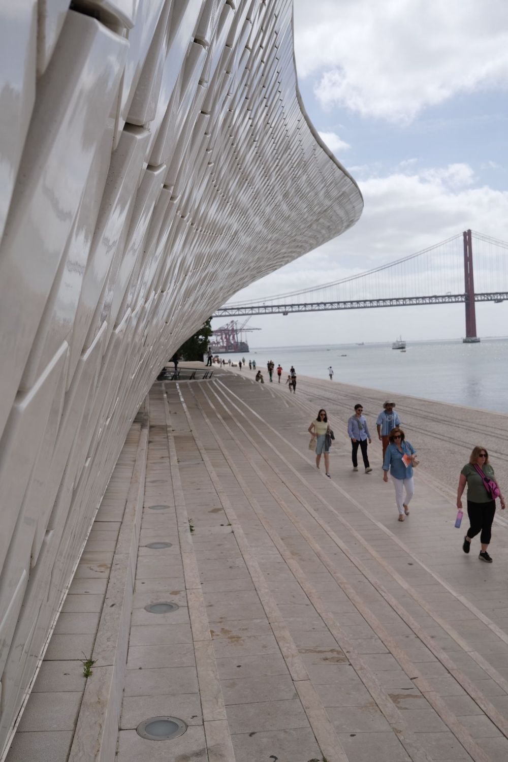 Modern MAAT museum facade in Lisbon with wooden promenade, visitors, and Ponte 25 de Abril bridge over Tagus River.