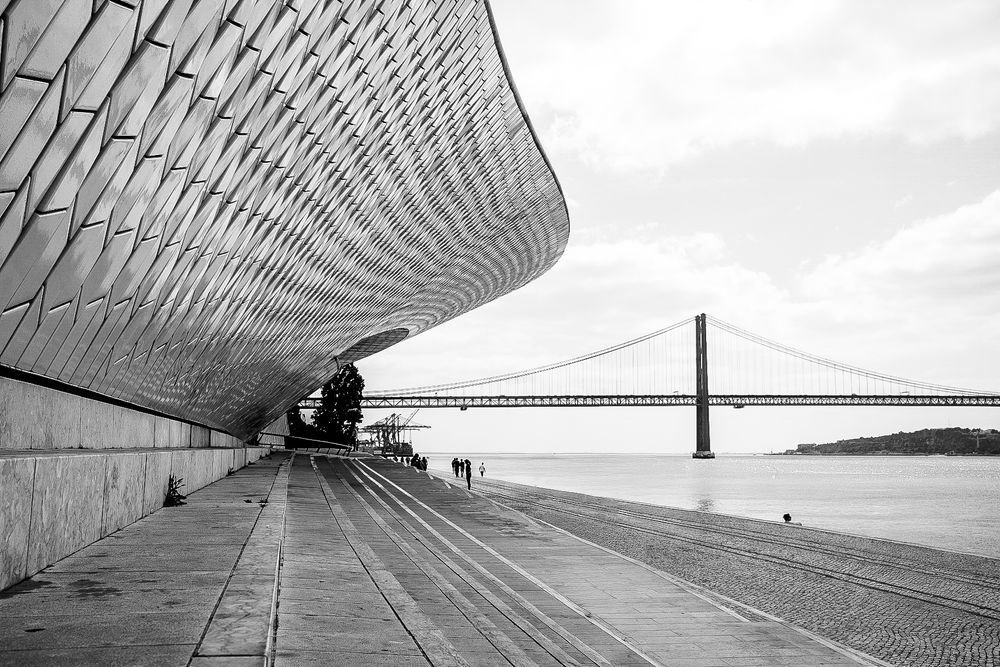Modern MAAT museum facade in Lisbon with wooden promenade, visitors, and Ponte 25 de Abril bridge over Tagus River.