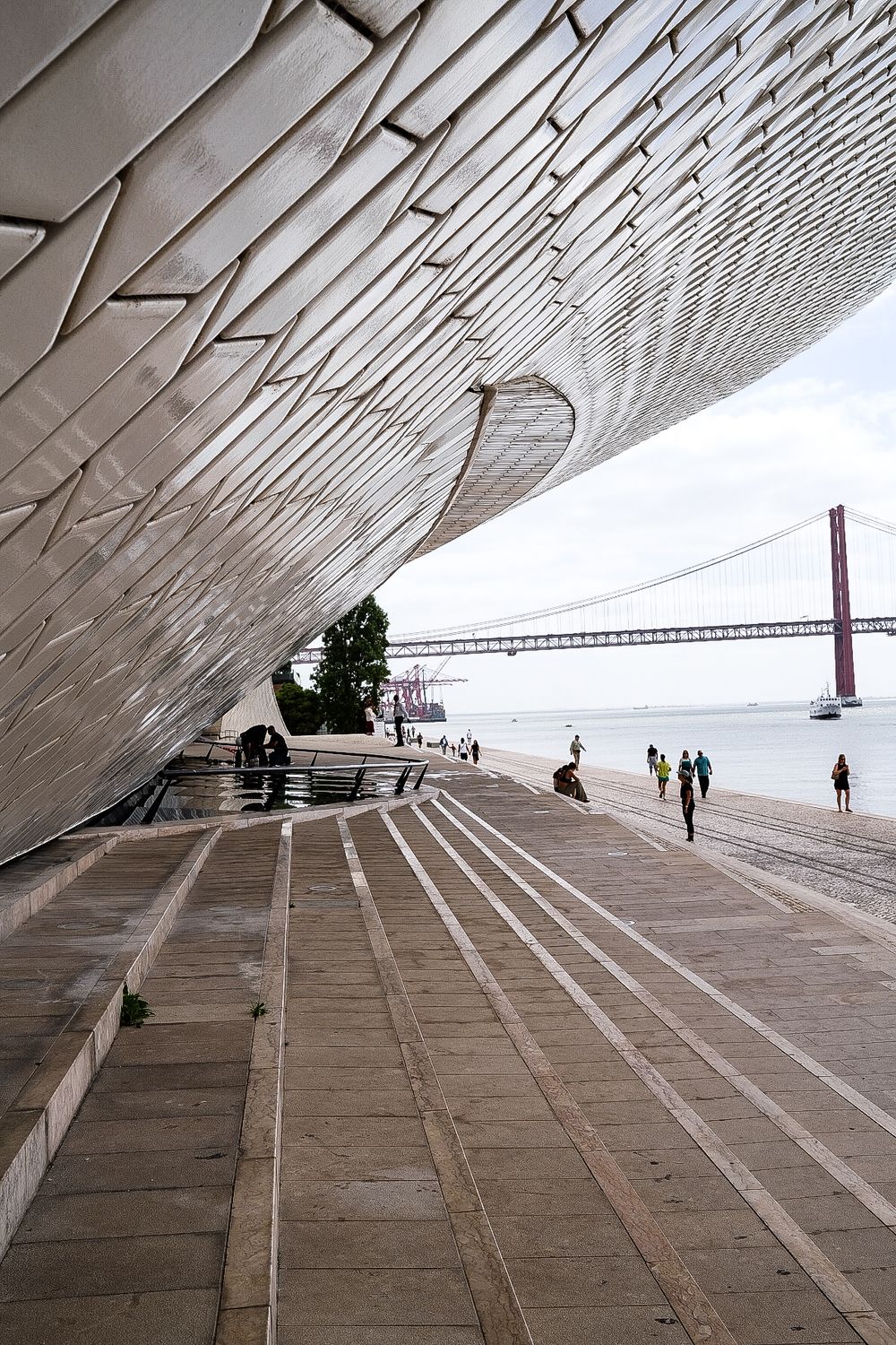 Modern MAAT museum facade in Lisbon with wooden promenade, visitors, and Ponte 25 de Abril bridge over Tagus River.