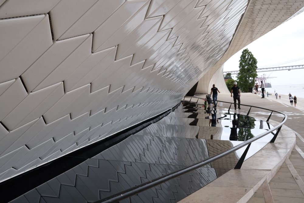Modern MAAT museum facade in Lisbon with wooden promenade, visitors, and Ponte 25 de Abril bridge over Tagus River.