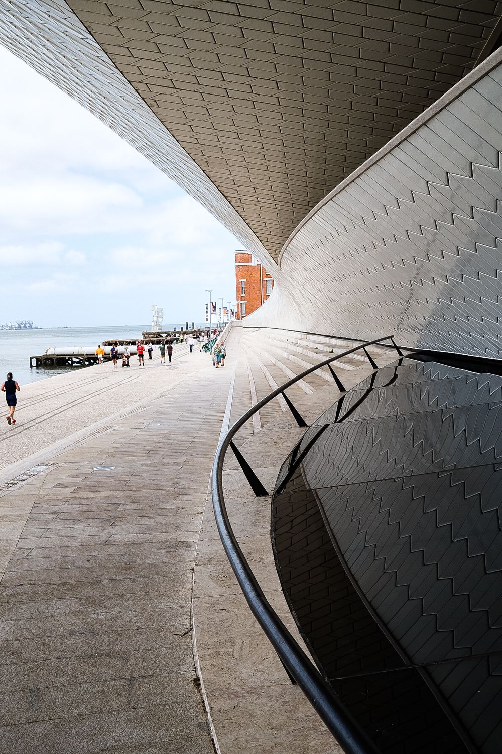Modern MAAT museum facade in Lisbon with wooden promenade, visitors, and Ponte 25 de Abril bridge over Tagus River.