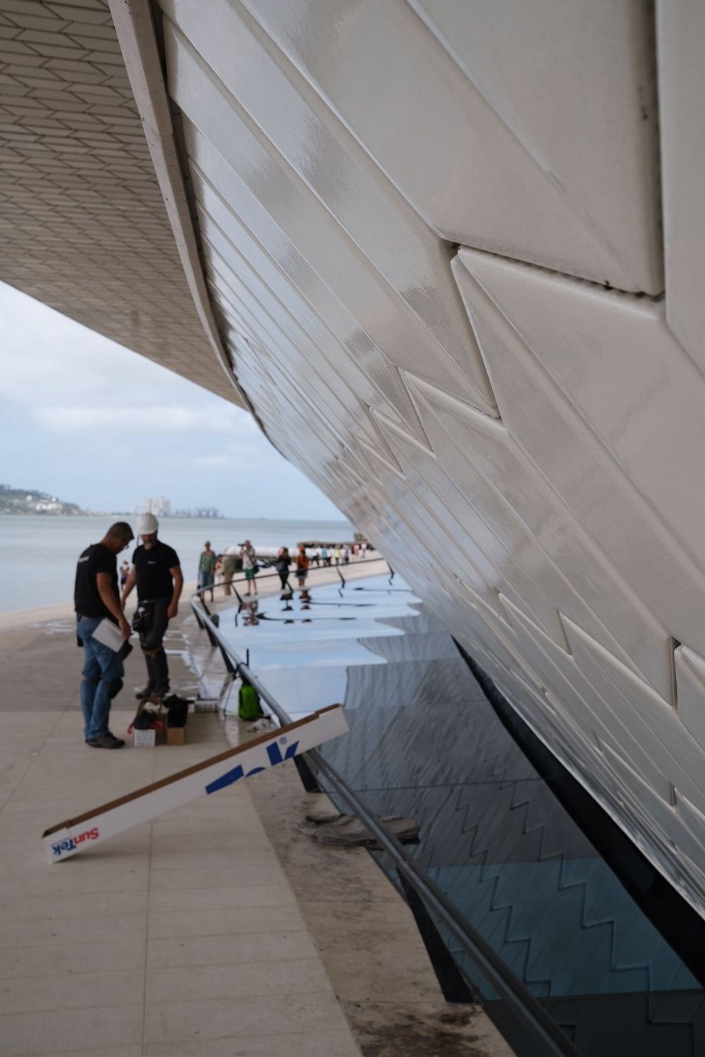Modern MAAT museum facade in Lisbon with wooden promenade, visitors, and Ponte 25 de Abril bridge over Tagus River.