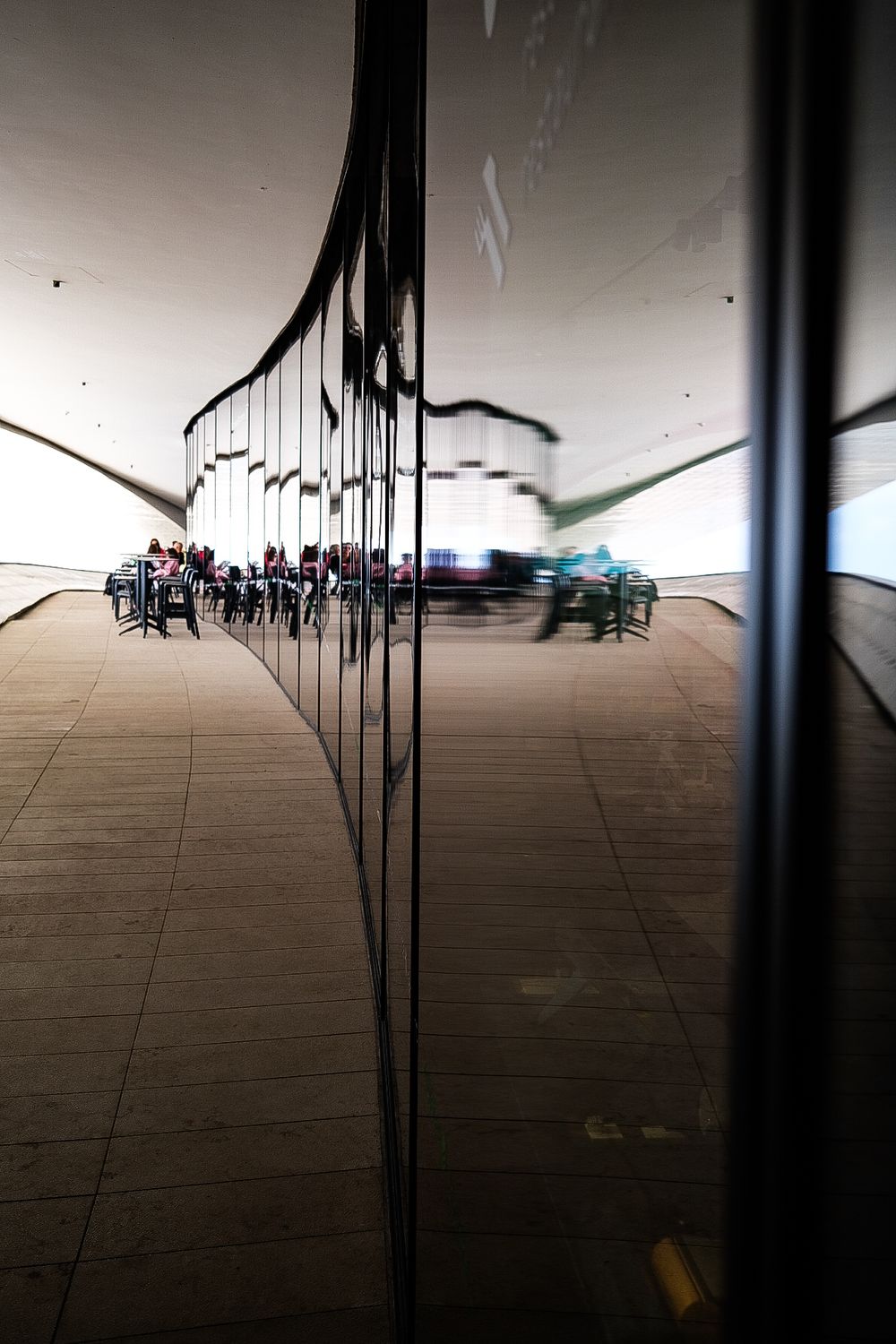 Modern MAAT museum facade in Lisbon with wooden promenade, visitors, and Ponte 25 de Abril bridge over Tagus River.