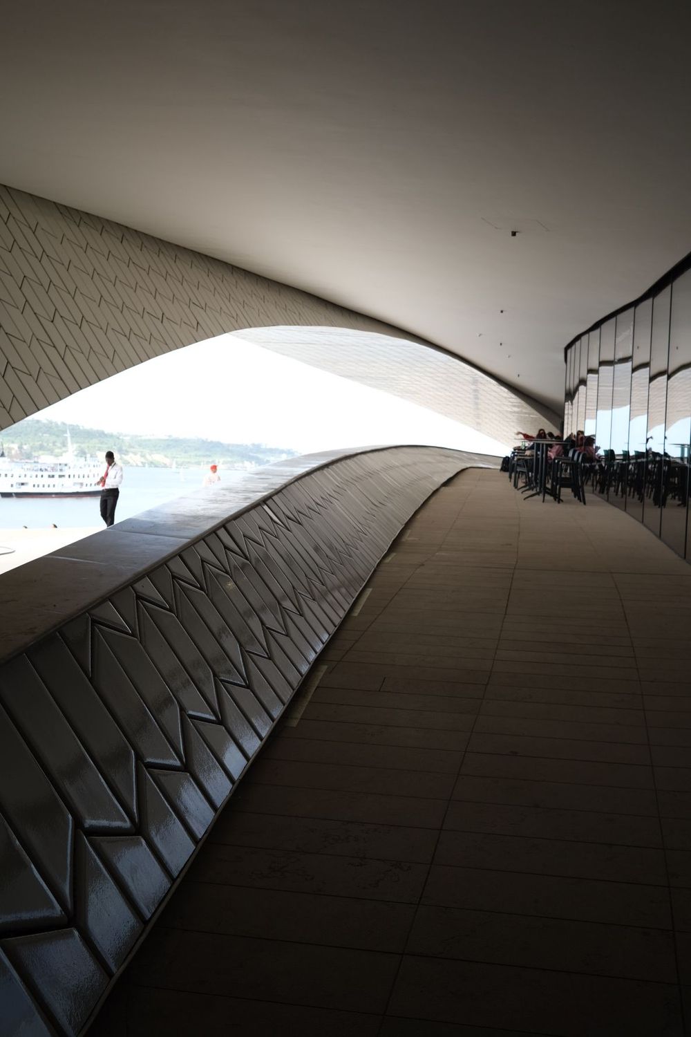 Modern MAAT museum facade in Lisbon with wooden promenade, visitors, and Ponte 25 de Abril bridge over Tagus River.