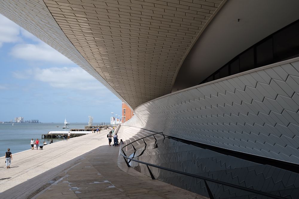 Modern MAAT museum facade in Lisbon with wooden promenade, visitors, and Ponte 25 de Abril bridge over Tagus River.