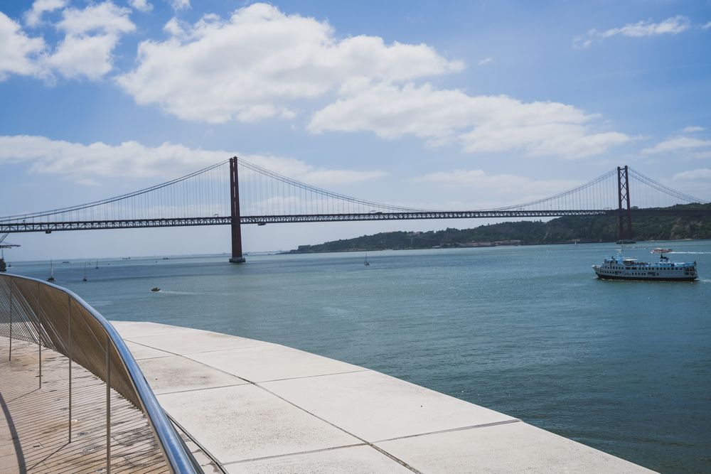 Modern MAAT museum facade in Lisbon with wooden promenade, visitors, and Ponte 25 de Abril bridge over Tagus River.