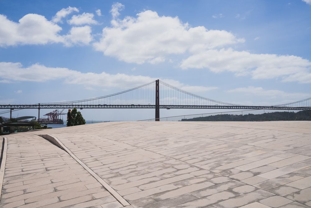 Modern MAAT museum facade in Lisbon with wooden promenade, visitors, and Ponte 25 de Abril bridge over Tagus River.
