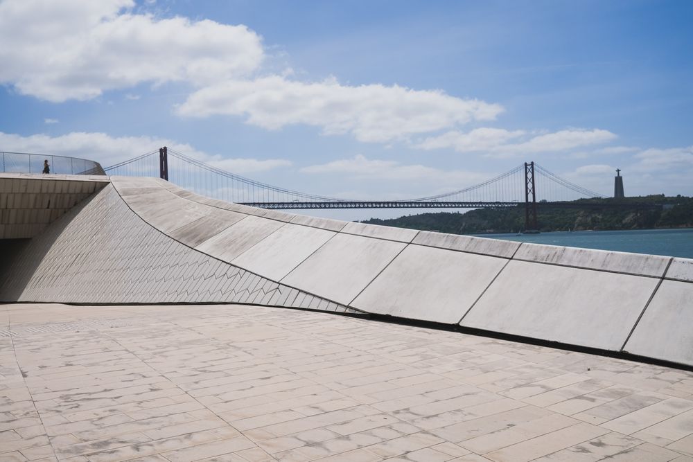 Modern MAAT museum facade in Lisbon with wooden promenade, visitors, and Ponte 25 de Abril bridge over Tagus River.
