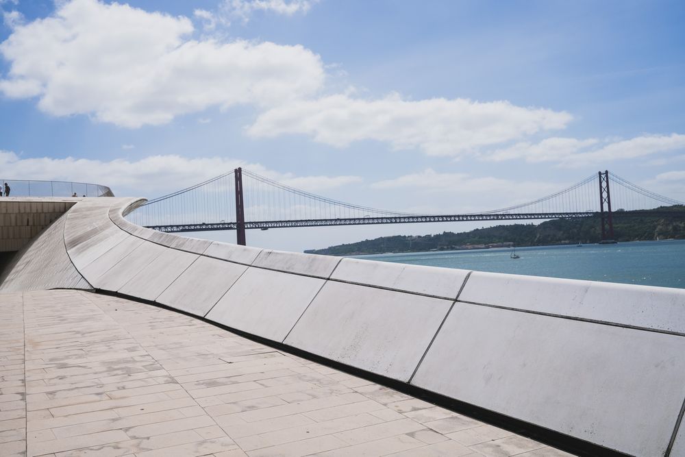 Modern MAAT museum facade in Lisbon with wooden promenade, visitors, and Ponte 25 de Abril bridge over Tagus River.