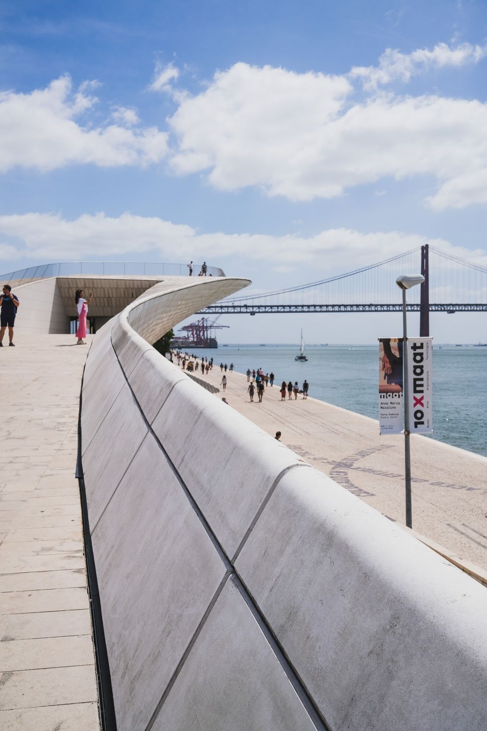 Modern MAAT museum facade in Lisbon with wooden promenade, visitors, and Ponte 25 de Abril bridge over Tagus River.