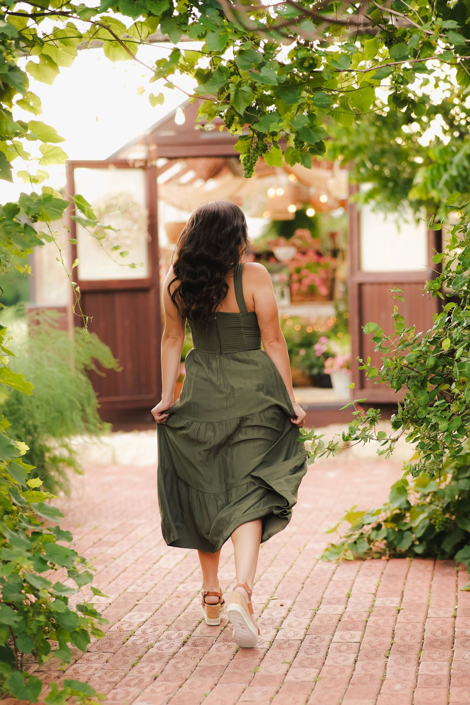 Woman in olive green dress walking away down brick path toward greenhouse under leafy green arch.