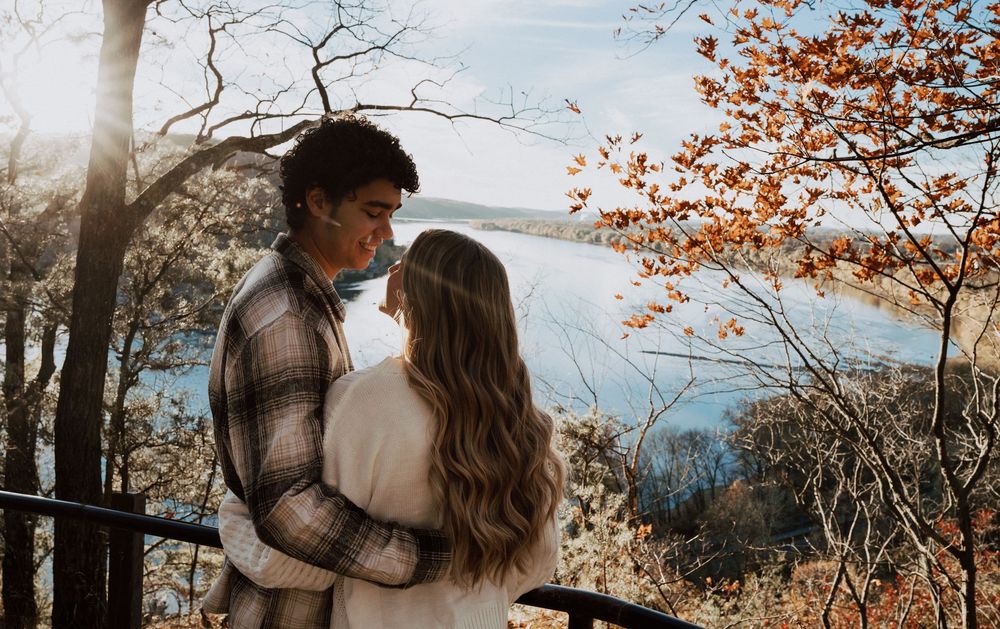 Couple embracing on a scenic overlook with autumn foliage and a serene lake in the background on a sunny day.