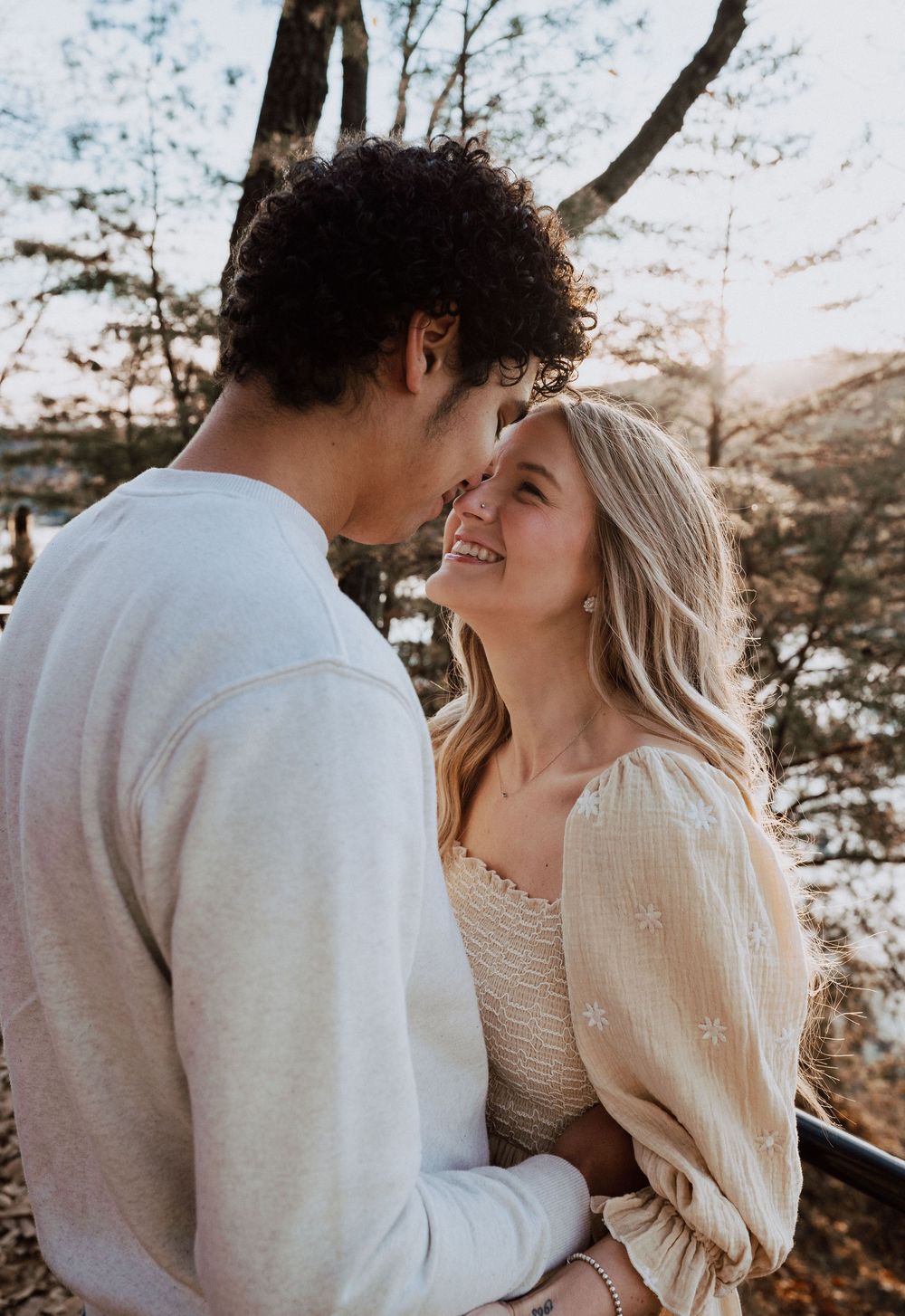 A smiling blonde woman and curly-haired man share a romantic moment outdoors in warm autumn light.