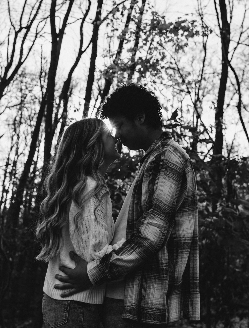 Black and white photo of a romantic couple almost kissing outdoors among bare trees in a forest setting.