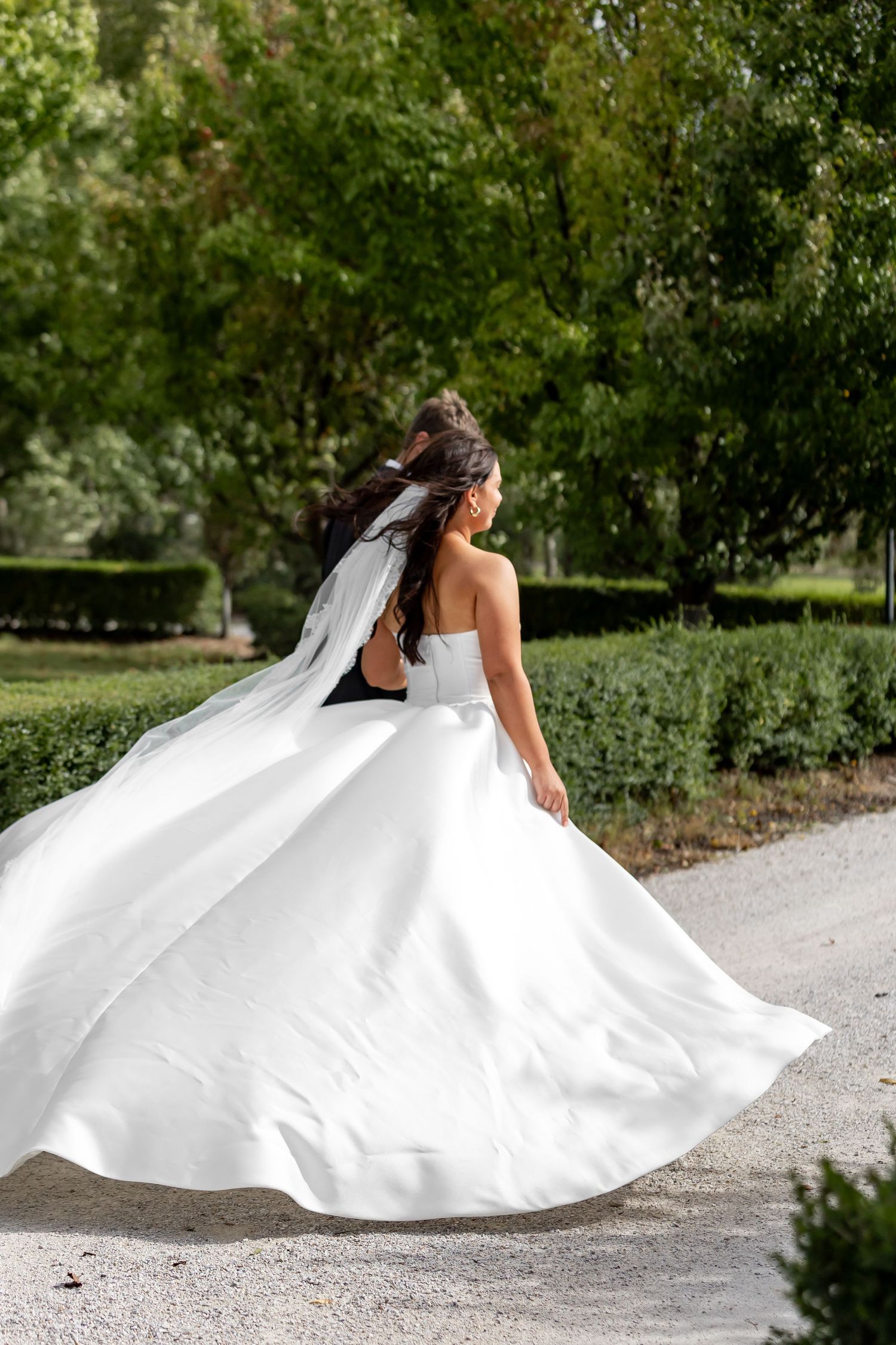 Bride in flowing white ball gown and veil twirls on garden path surrounded by lush green hedges.