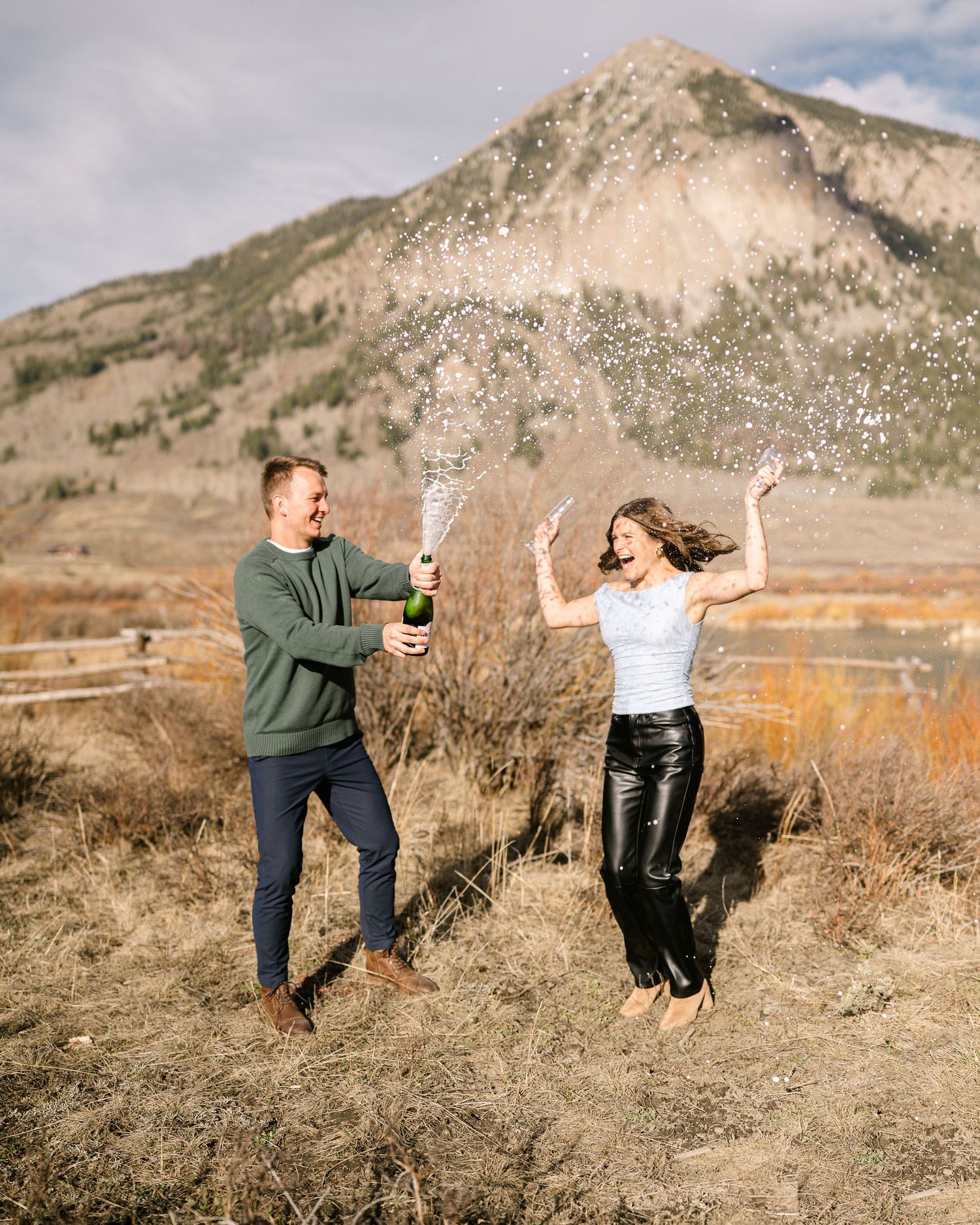 Couple celebrating engagement by popping champagne bottle outdoors with mountain backdrop in Colorado.