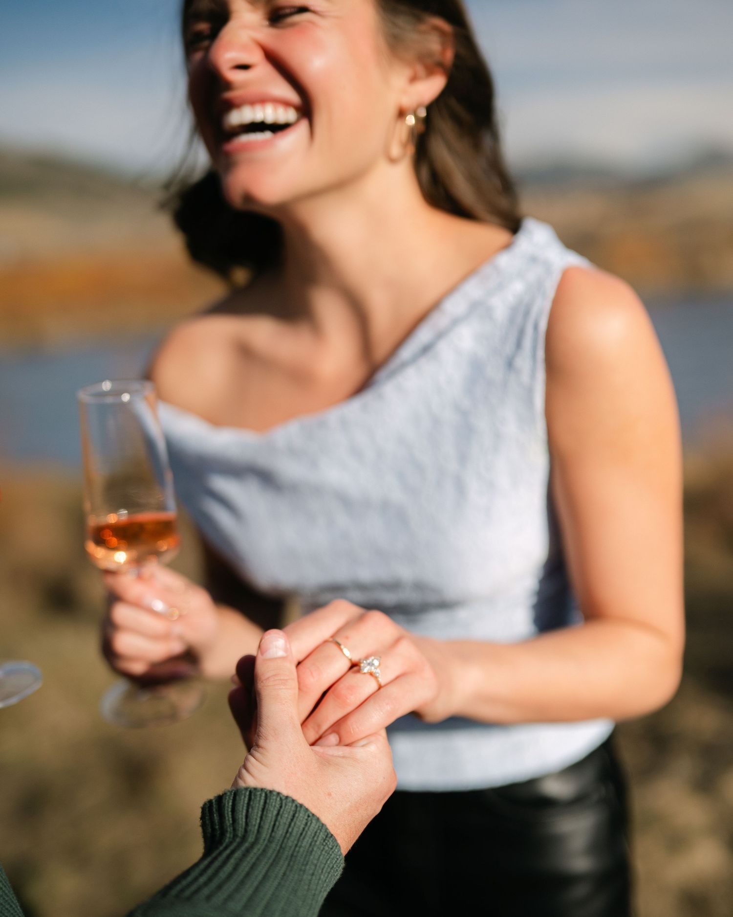 Joyful woman laughing holding champagne glass while showing off engagement ring outdoors.