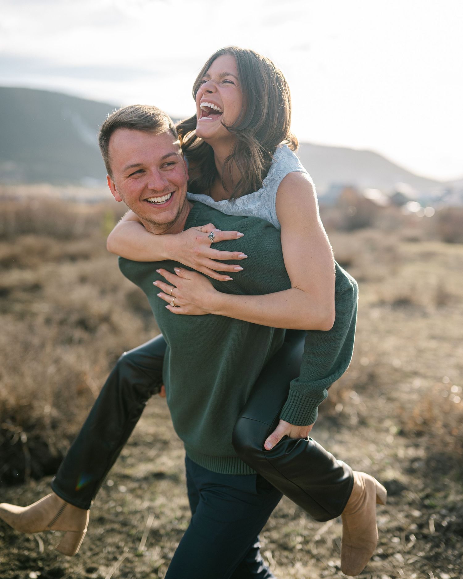 A happy couple laughing outdoors, man giving woman a piggyback ride in a scenic field.