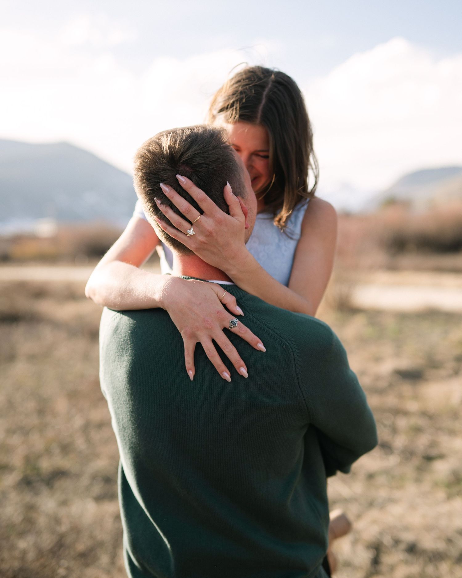 Couple sharing a tender embrace outdoors with mountains in the background, woman showing engagement ring.
