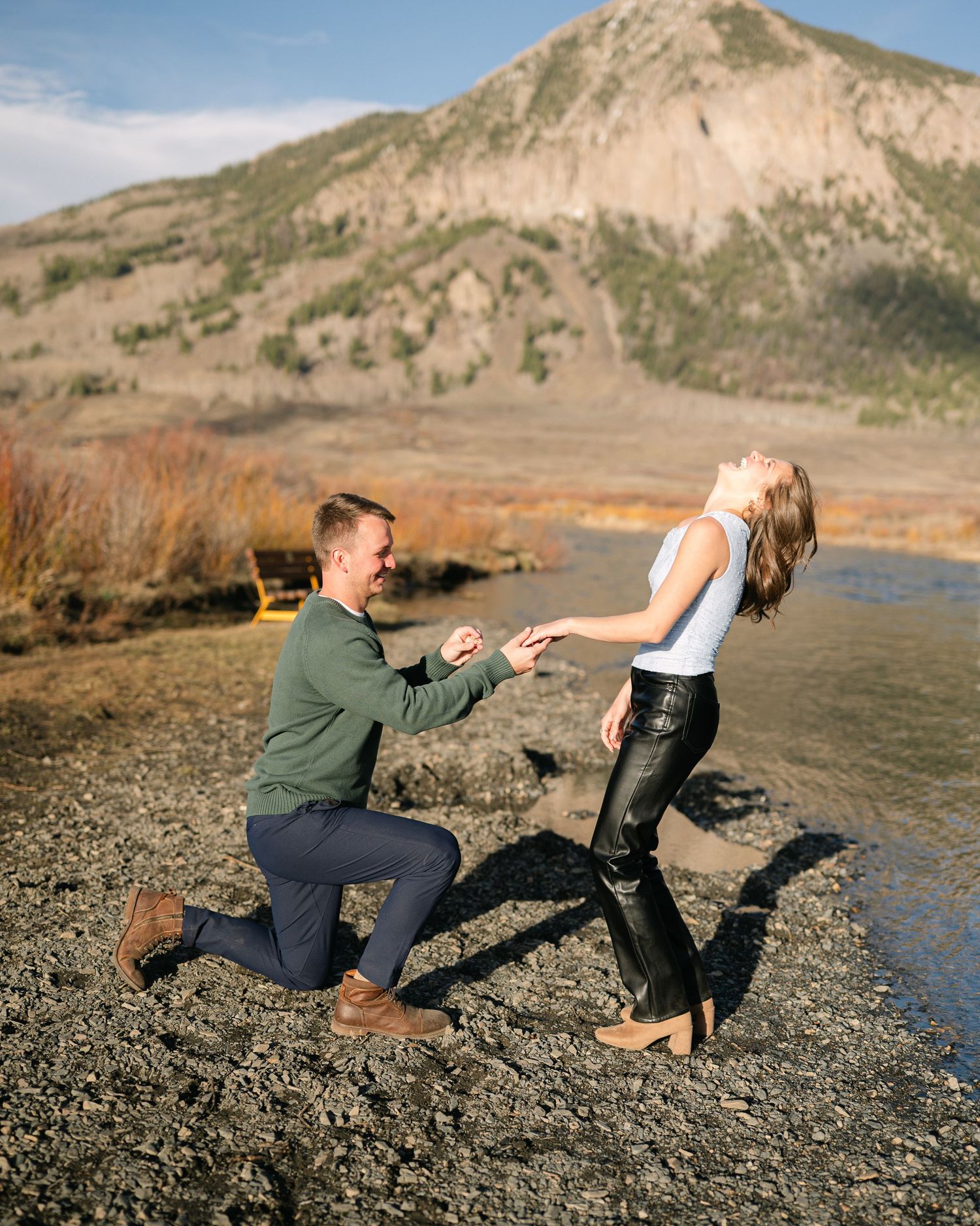Man proposing on one knee to a laughing woman by a mountain lake in a scenic outdoor setting.