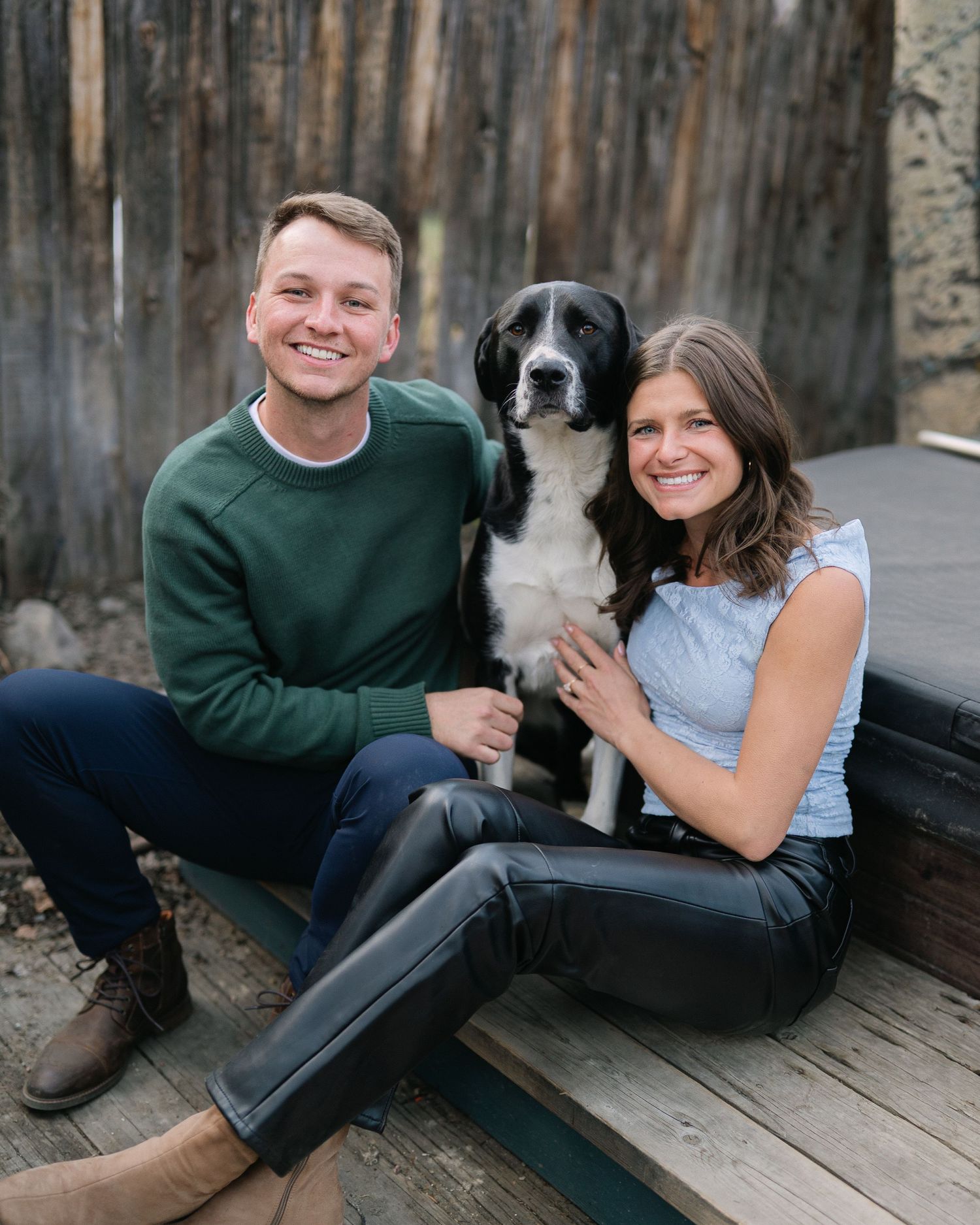 Smiling couple sitting on wooden steps with their black and white dog in a rustic outdoor setting.