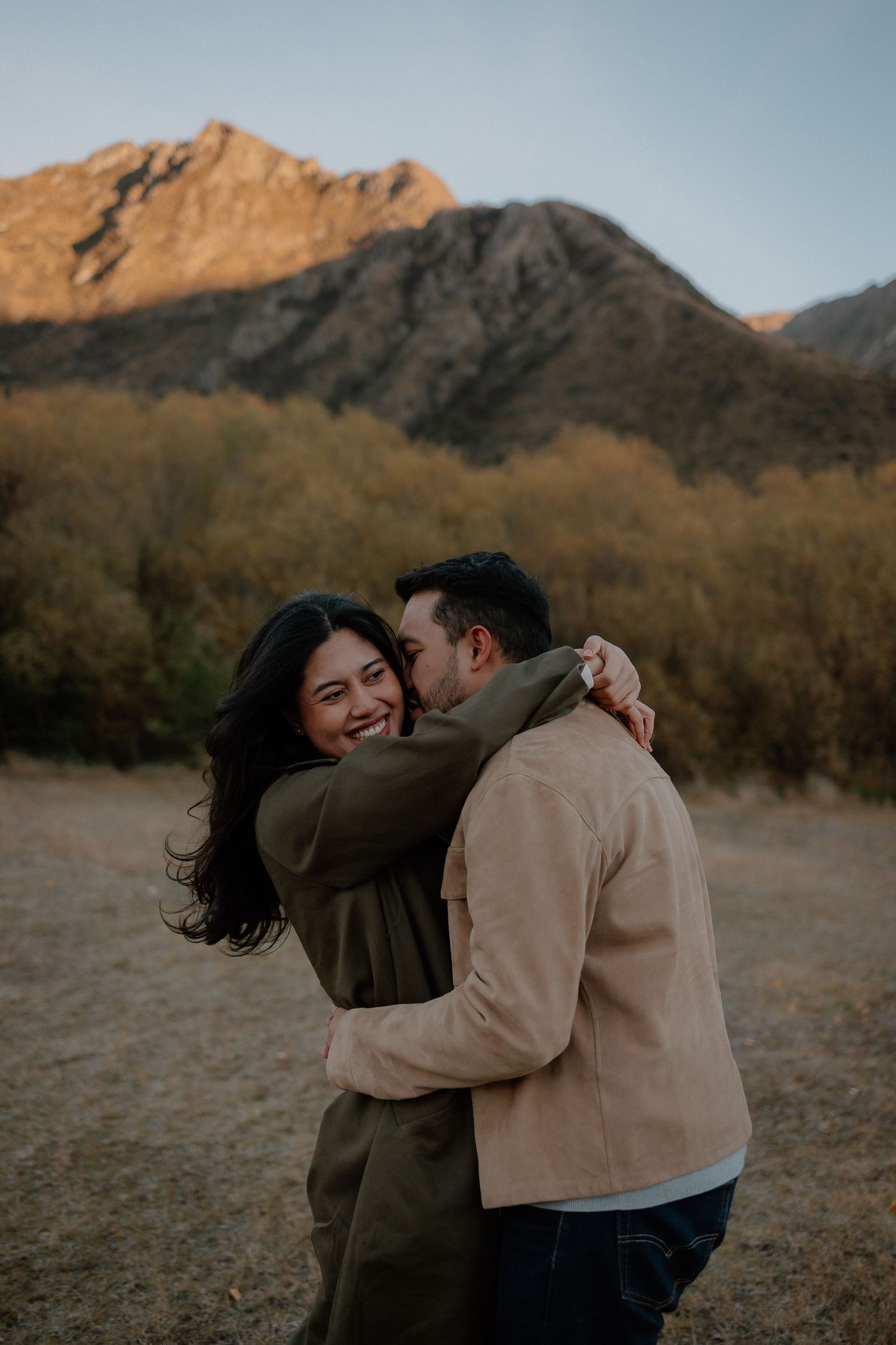 Couple embracing and laughing in a mountain field at sunset, woman in olive coat, man in tan jacket.