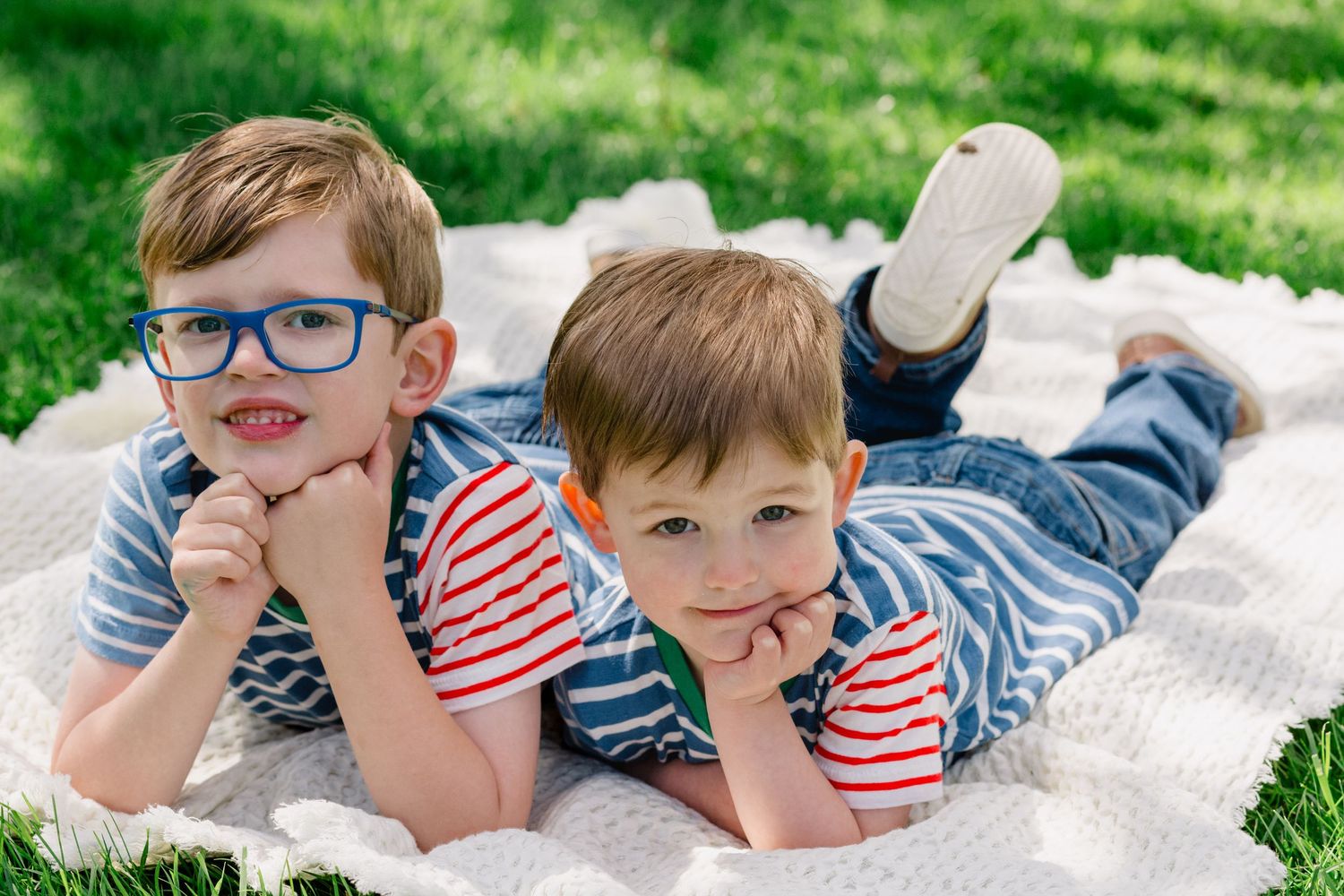 Two young boys in striped shirts lying on a white blanket in green grass, smiling at the camera.