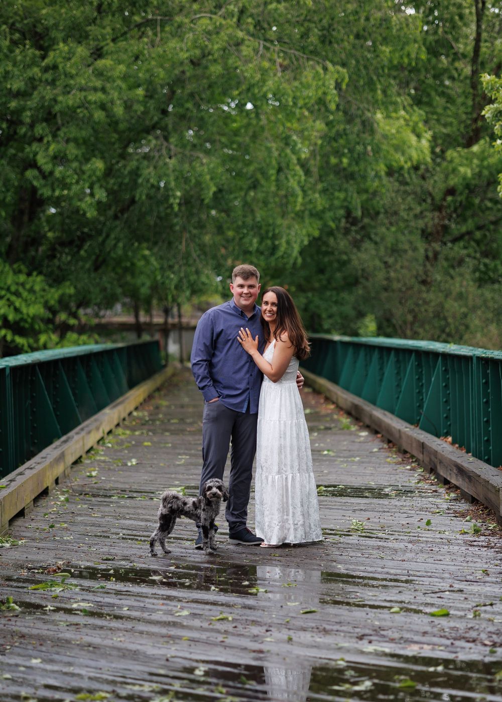 Couple poses with small dog on rainy wooden bridge surrounded by lush green trees.