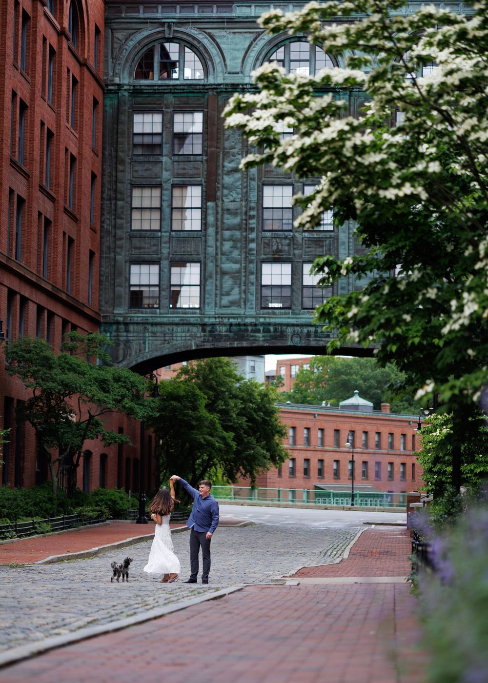 Couple in wedding attire with small black dog on cobblestone street between historic brick buildings with green bridge overhead.