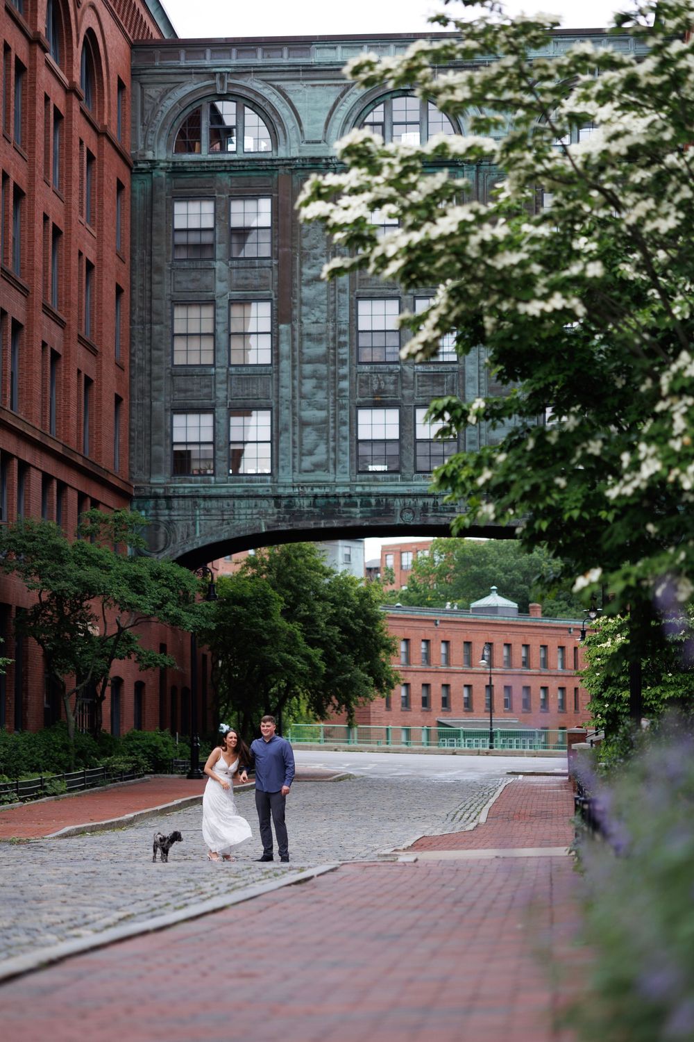 Bride and groom walking hand in hand on brick cobblestone path flanked by red brick buildings and green skybridge.
