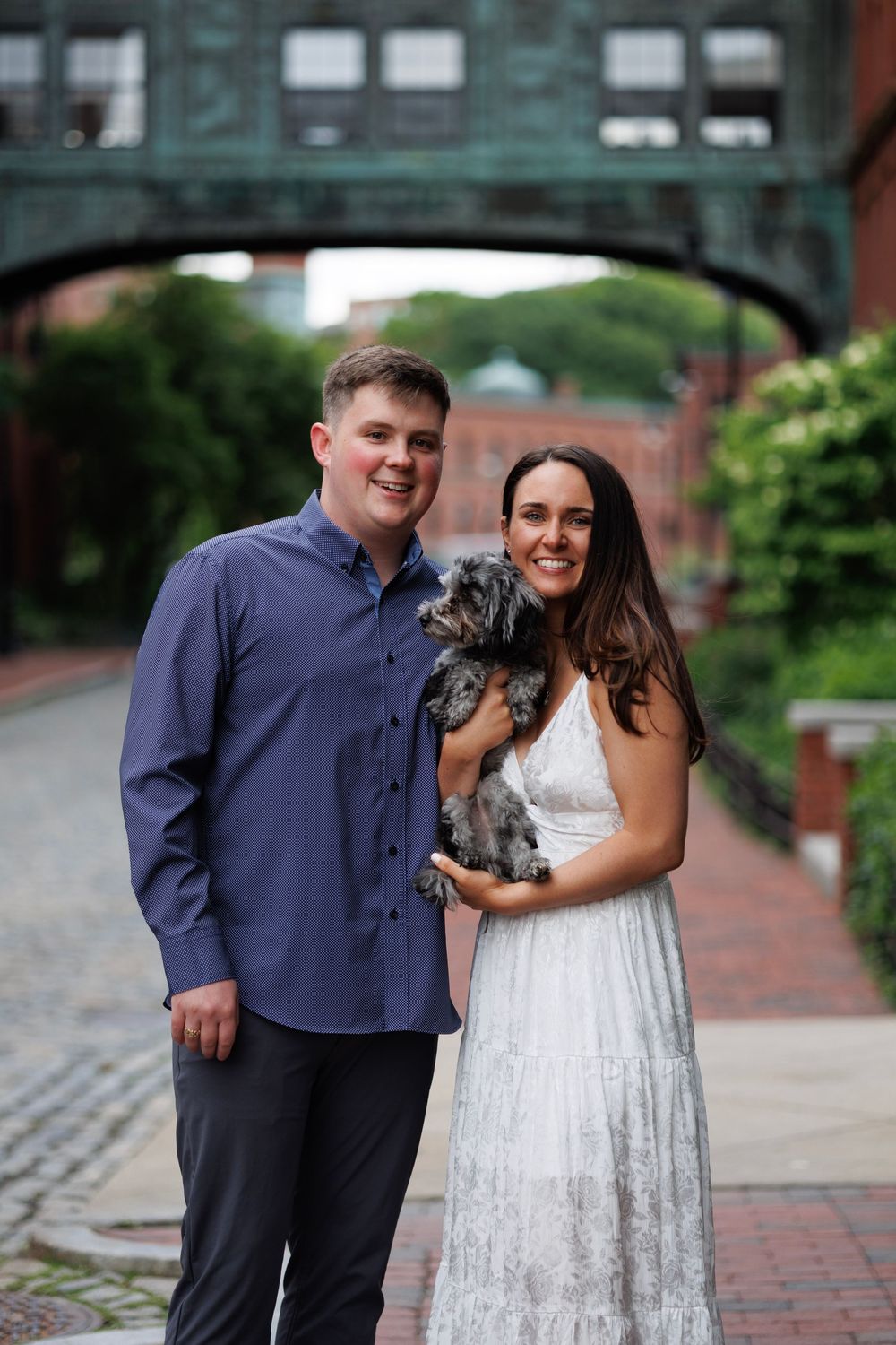 Smiling couple holds small gray dog in urban brick courtyard under green arch bridge.