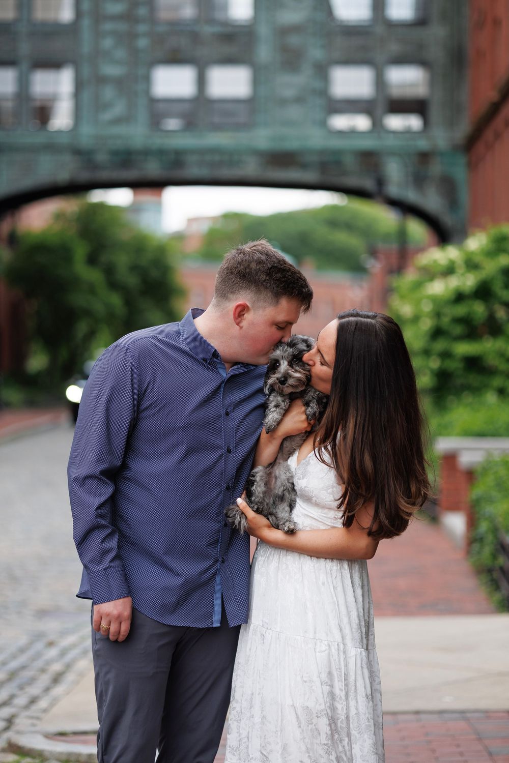 Couple sharing a kiss with their small dog on a cobblestone street with brick buildings and green archway.