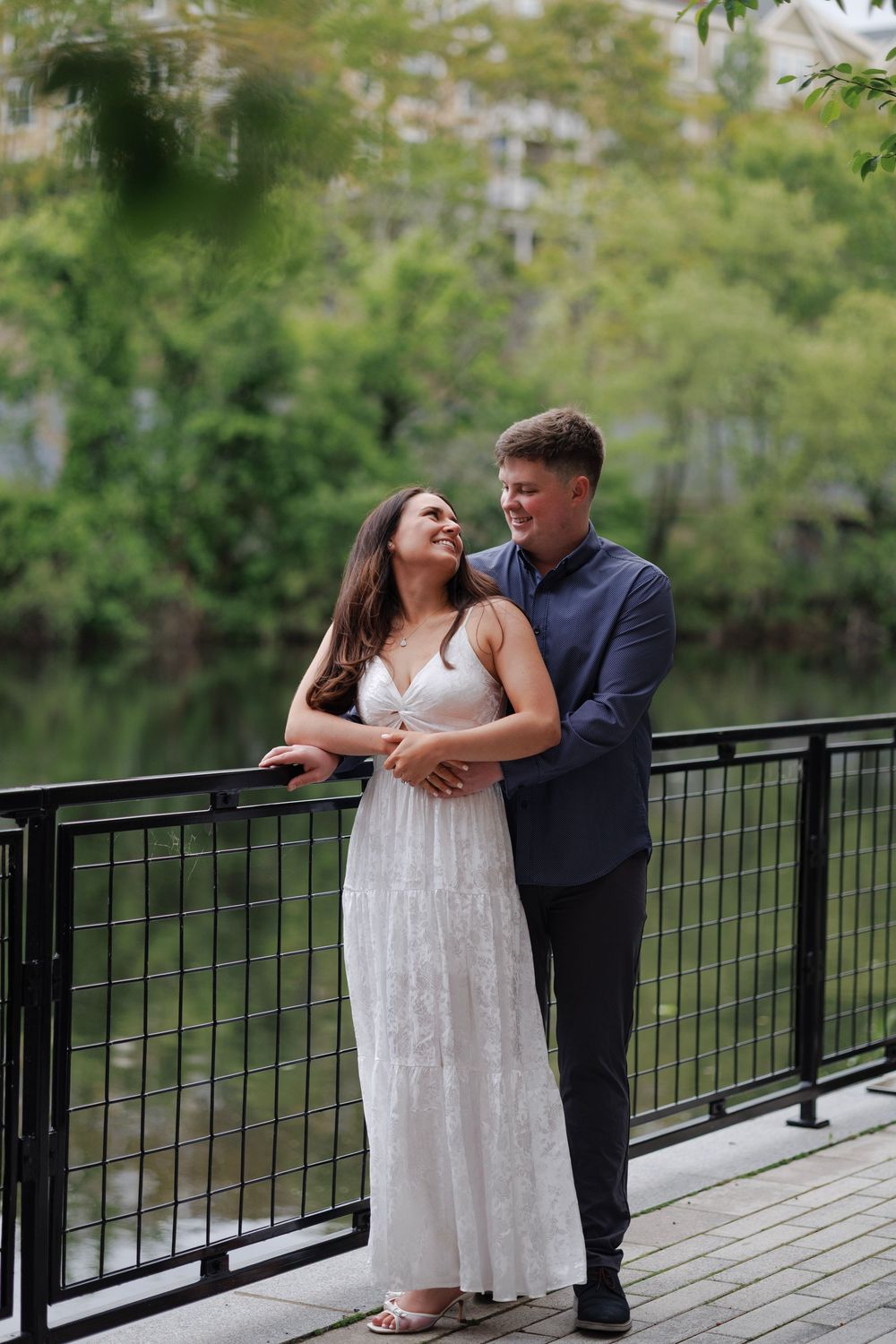 Couple embracing on a bridge surrounded by lush greenery, woman in white lace dress and man in navy shirt.