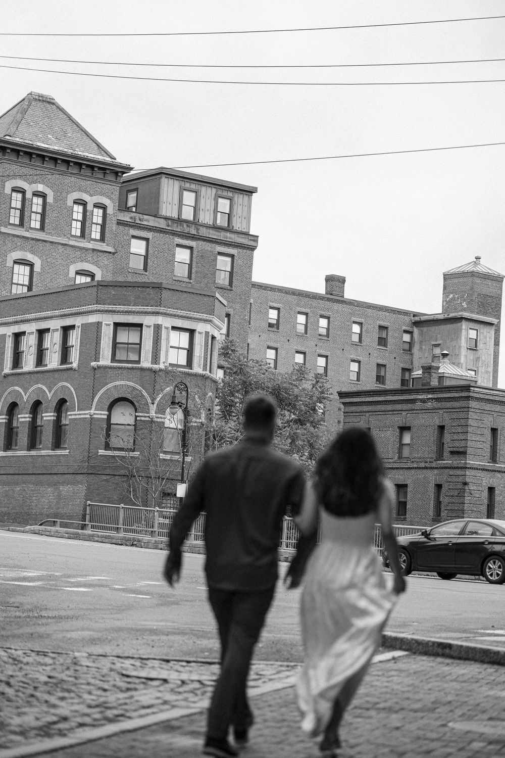Black and white photo of couple walking hand in hand on cobblestone street with historic brick buildings.