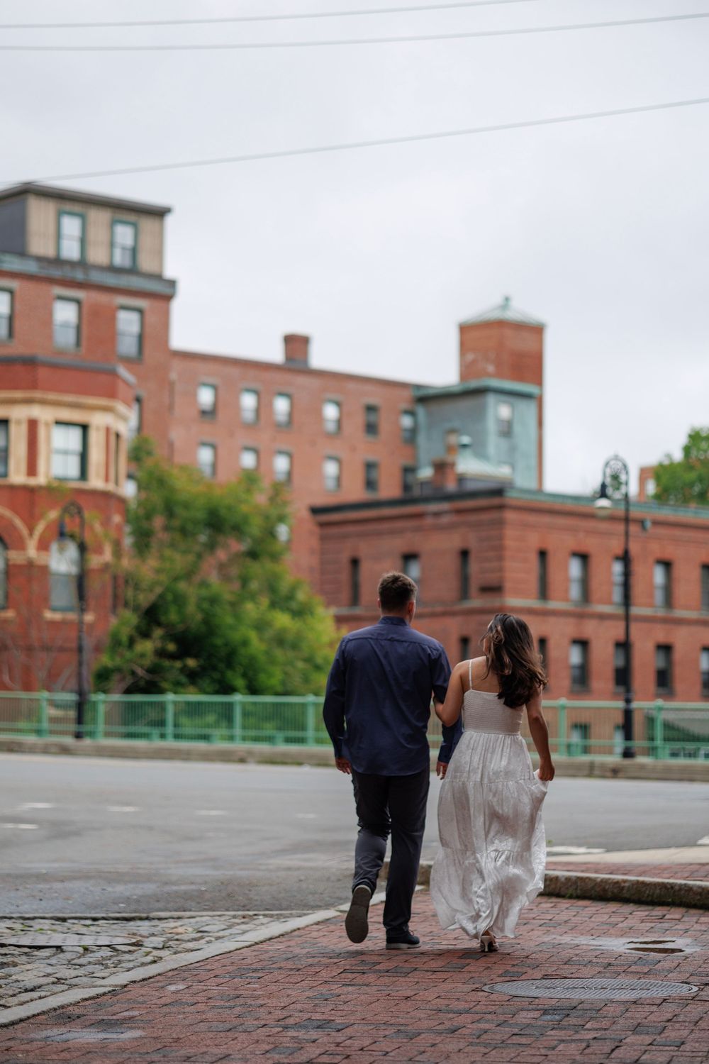 Couple holding hands walking away on brick path surrounded by historic red brick buildings on overcast day.
