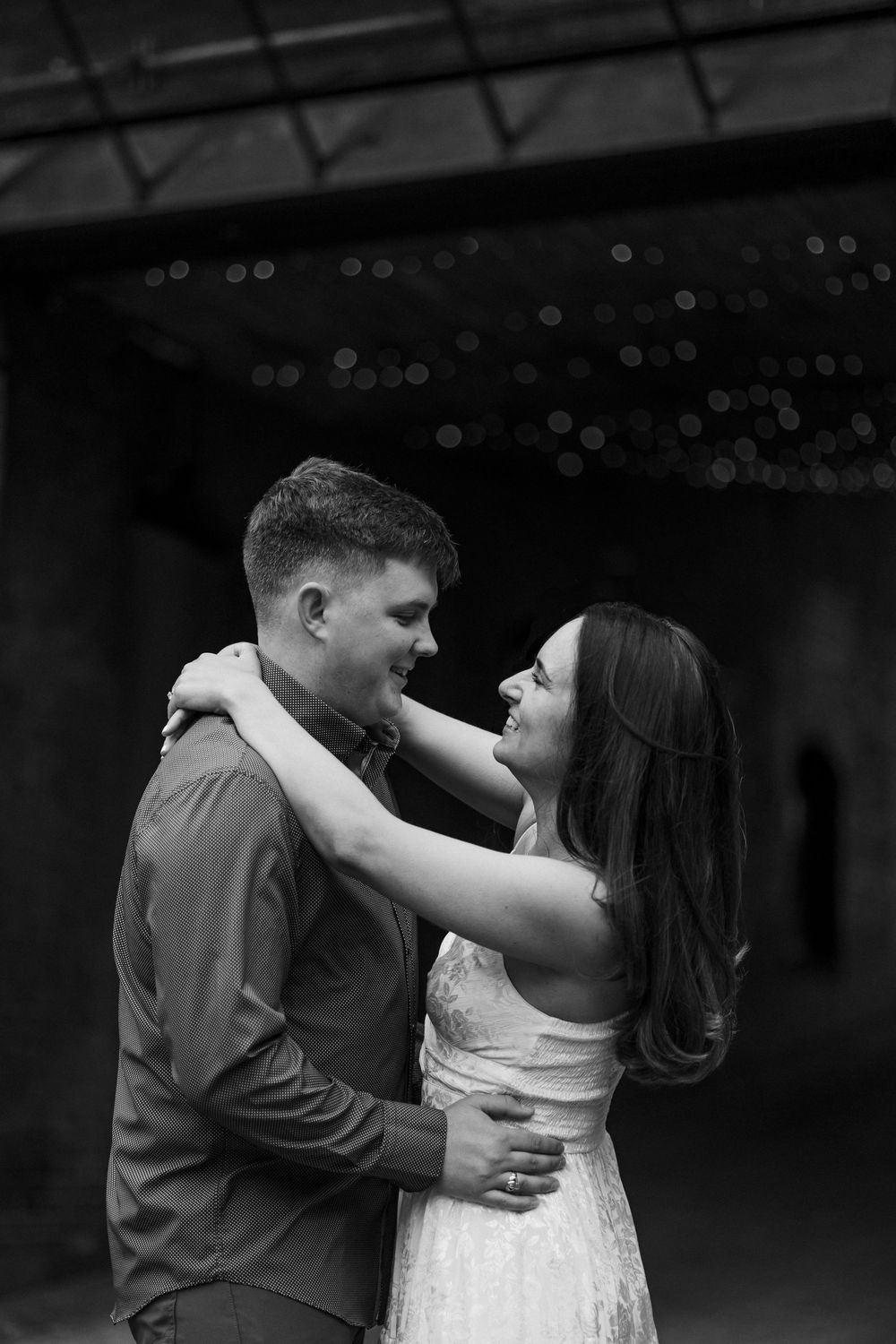 Black and white photo of a couple embracing and smiling at each other with bokeh lights in background.