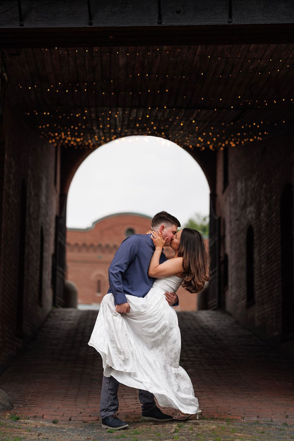 Couple sharing a romantic dip kiss under a brick archway adorned with warm string lights.
