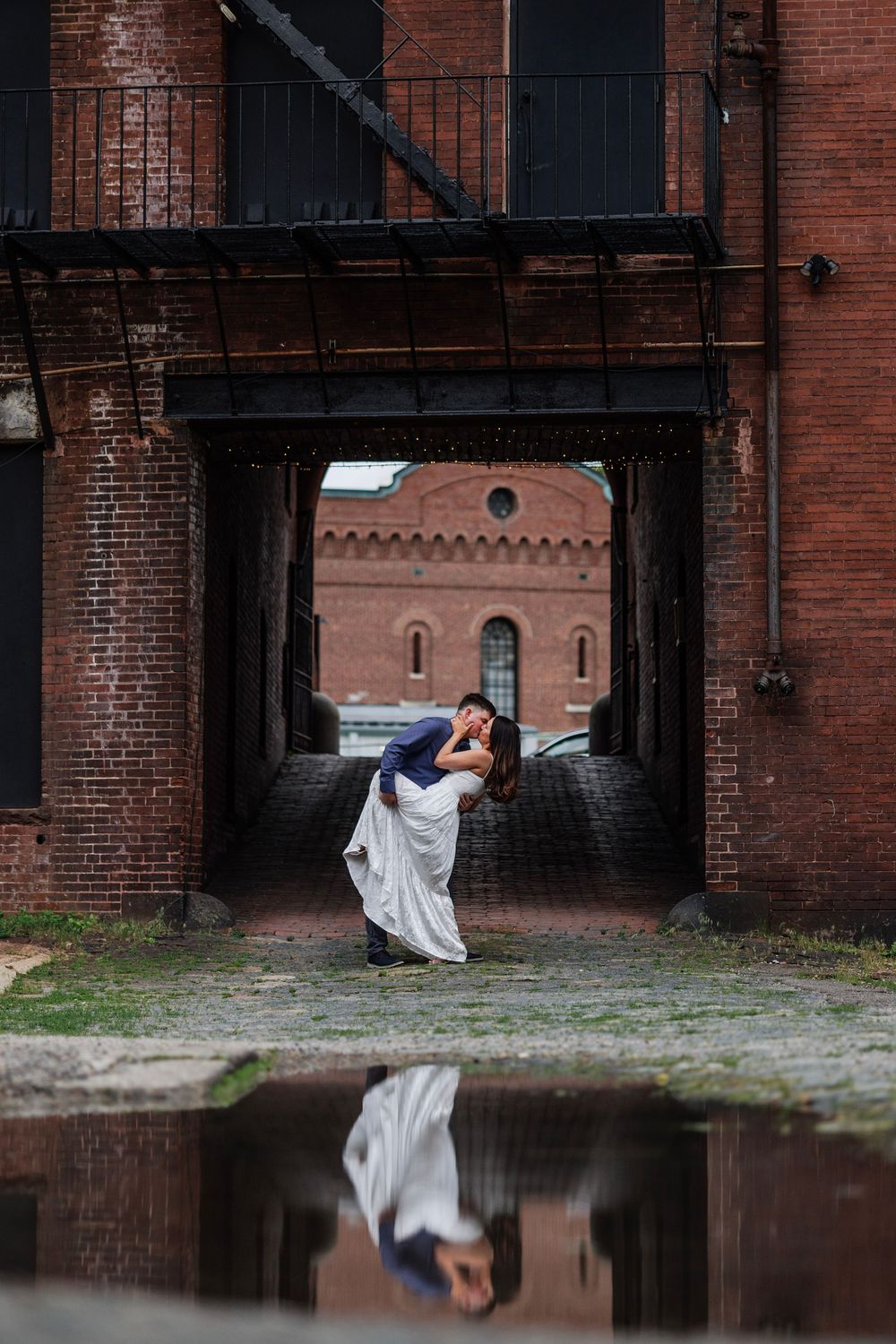 Couple sharing a dip kiss in a brick archway with their reflection visible in a puddle below.