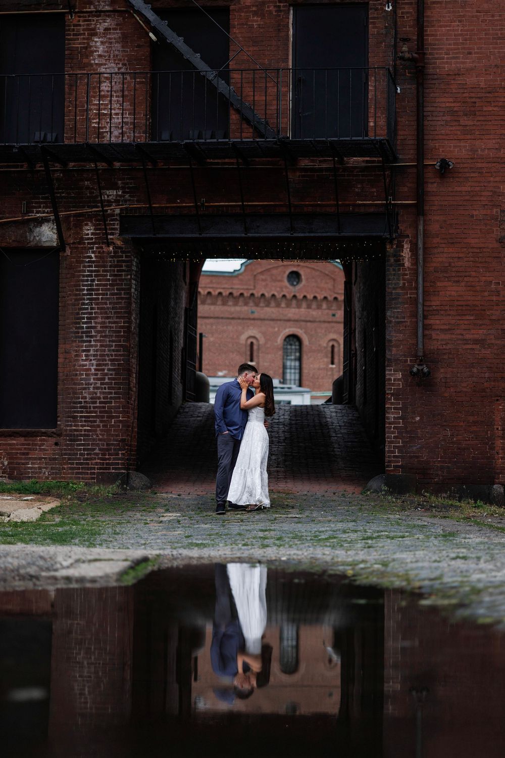 Couple sharing romantic kiss in brick archway with puddle reflection below in urban industrial setting.