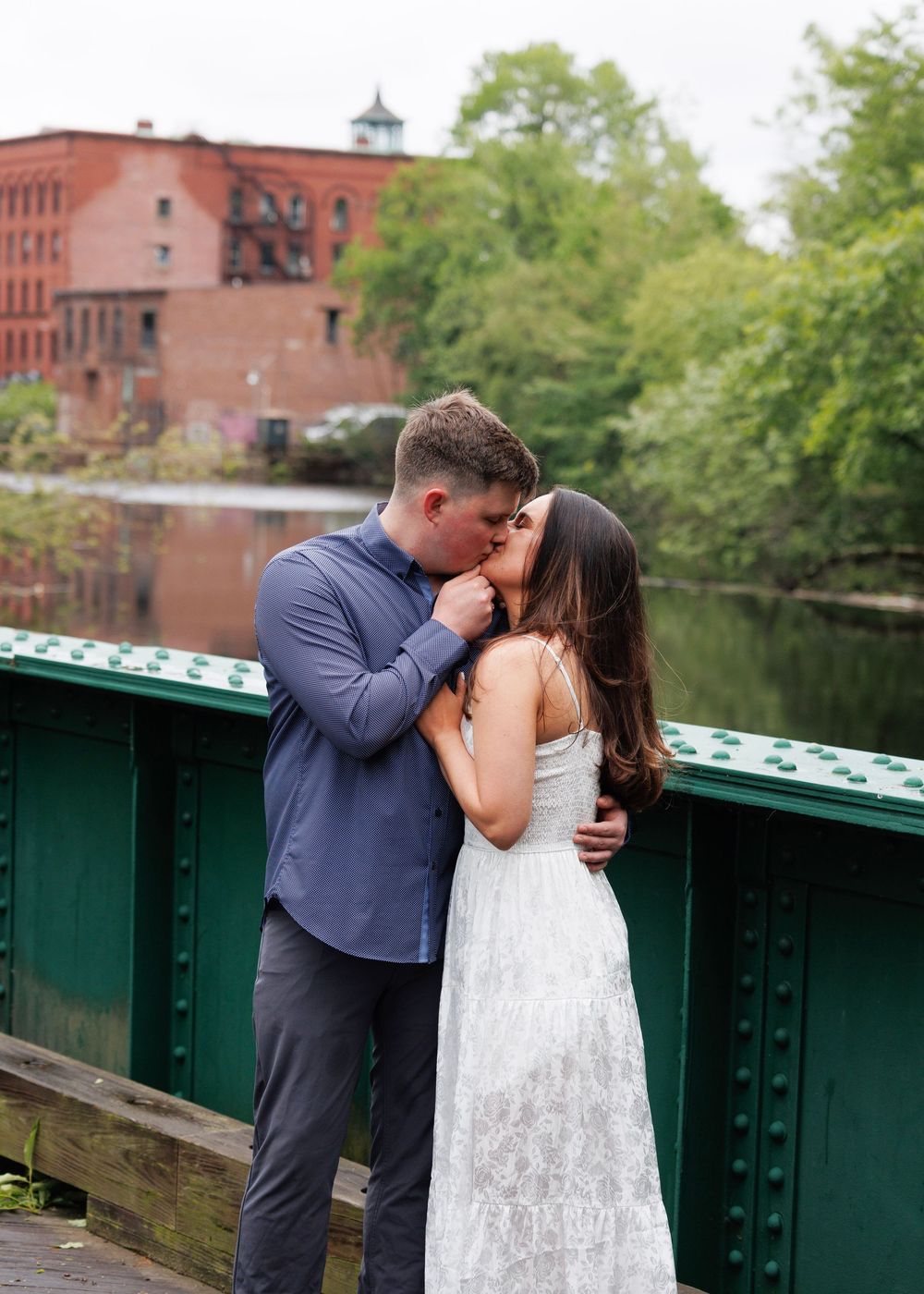 Couple sharing a kiss on a green iron bridge with a scenic river and brick buildings behind them.