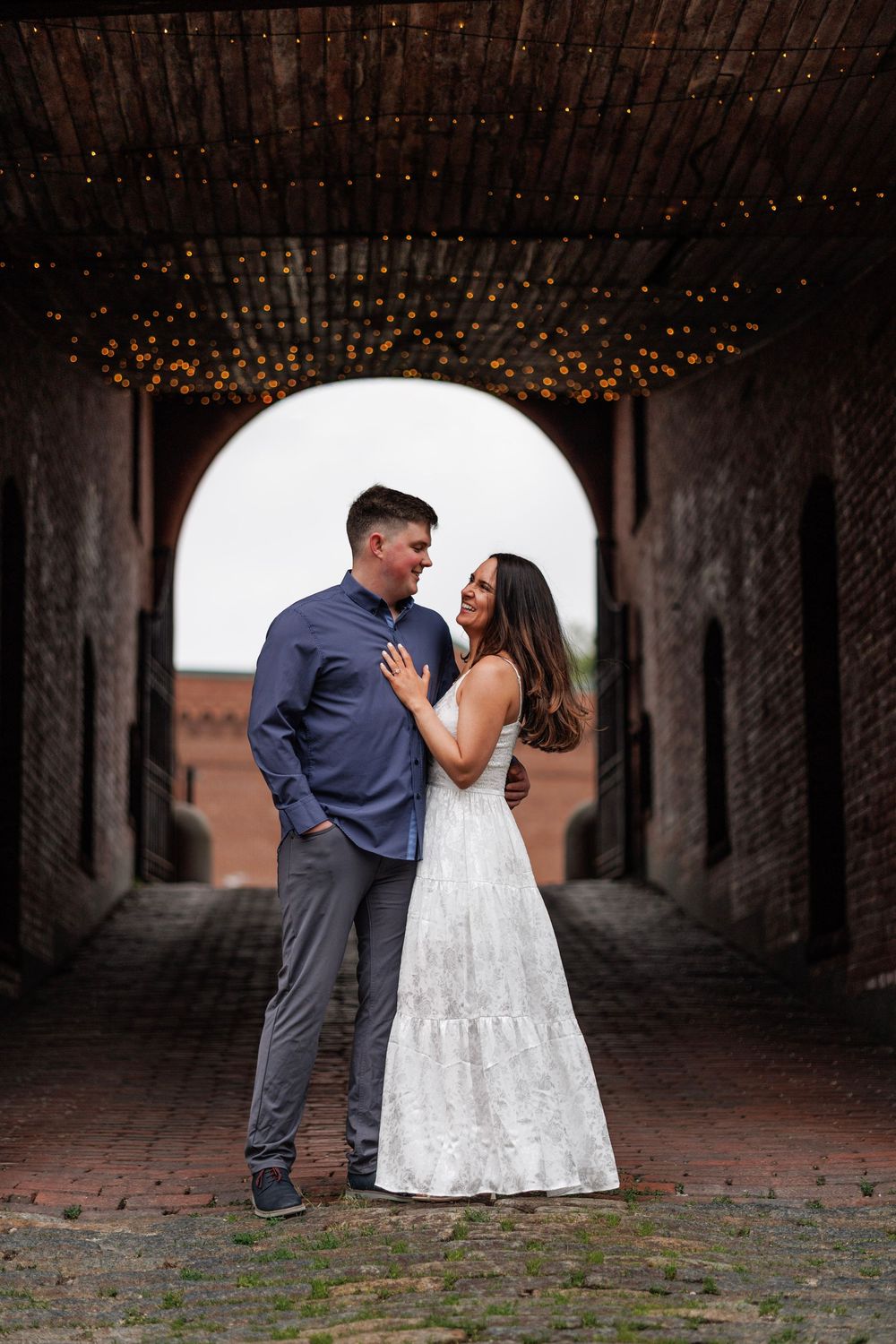 Couple in love posing under brick archway with string lights in white dress and blue shirt.