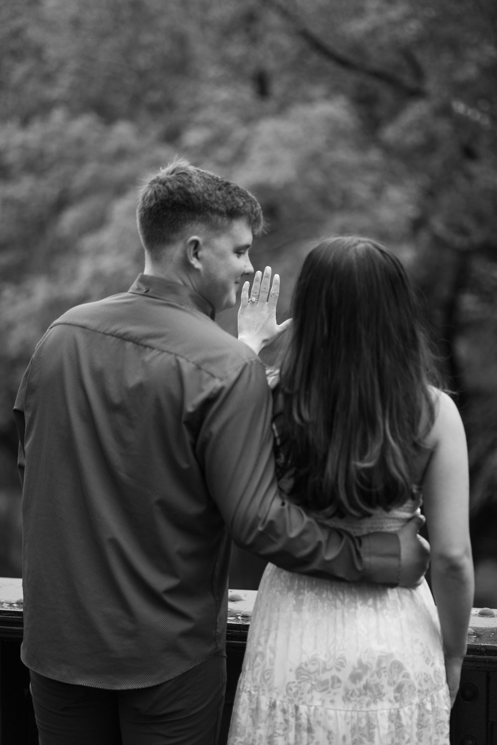 Black and white photo of a couple standing together on a bridge overlooking trees.