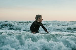 Child in black wetsuit playing in crashing ocean surf waves at sunset.