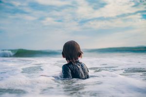 Child in wetsuit standing in ocean waves with cloudy sky backdrop.